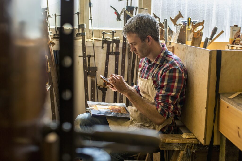 An antique furniture restorer sitting using a smart phone, surrounded by the tools of his trade