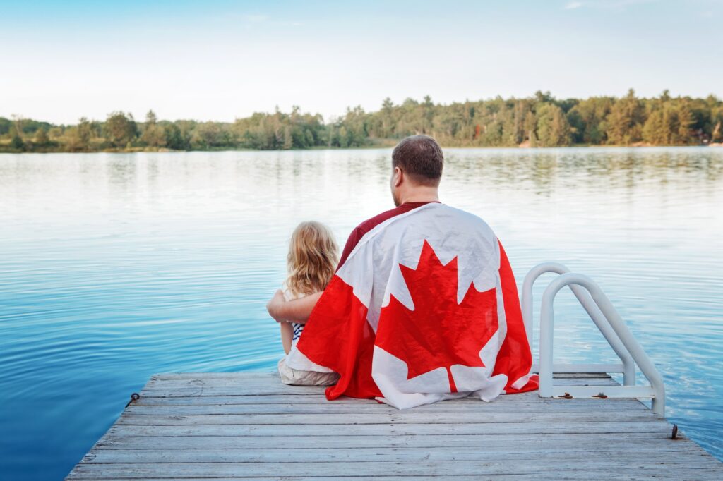 Father and daughter wrapped in large Canada flag sitting on pier by lake. Canada Day celebration.