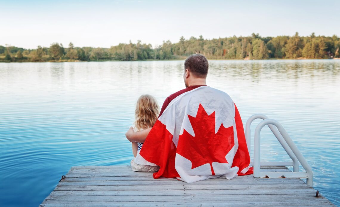 Father and daughter wrapped in large Canada flag sitting on pier by lake. Canada Day celebration.