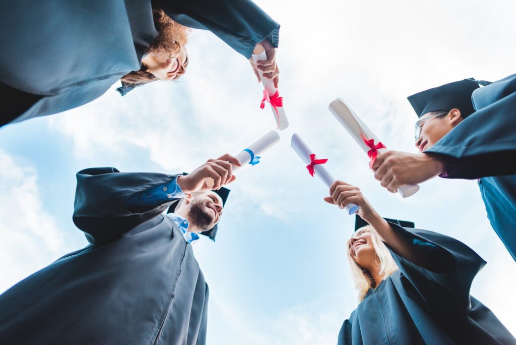 bottom view of multiracial graduates with diplomas in hands and blue sky on background