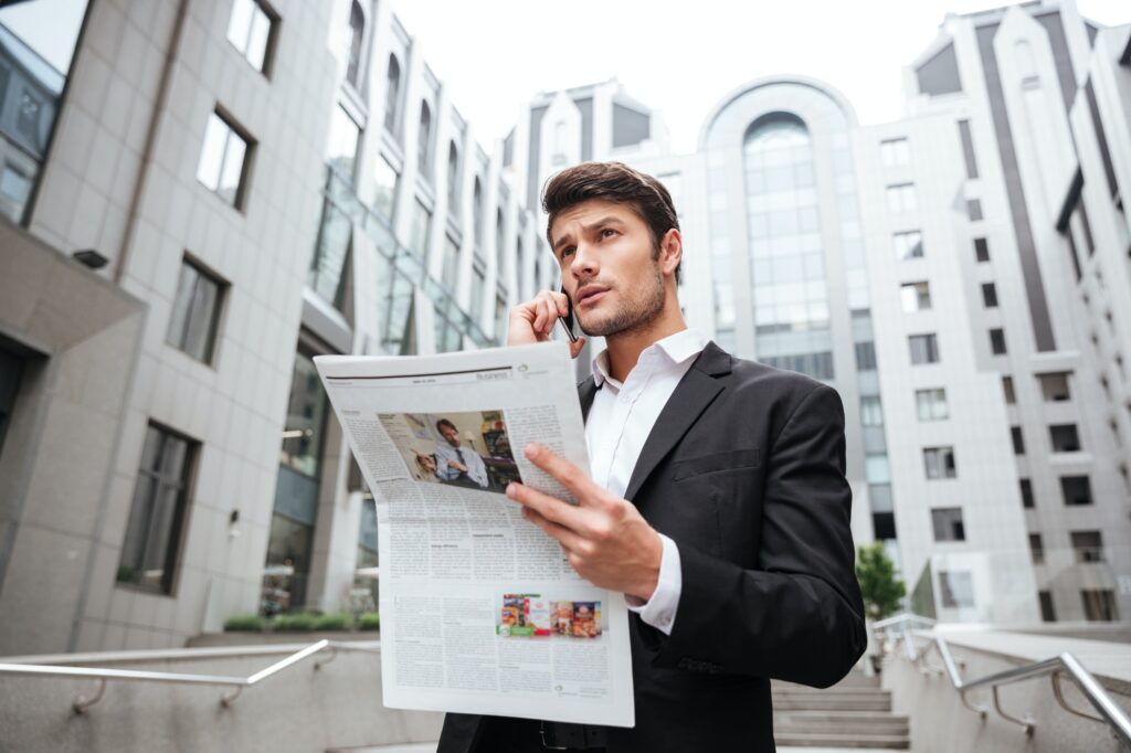 Pensive young businessman talking on mobile phone and reading newspaper
