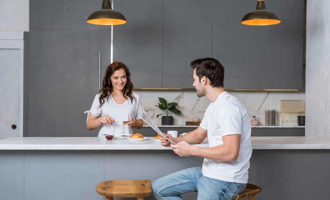beautiful girlfriend looking at handsome man holding newspaper in kitchen