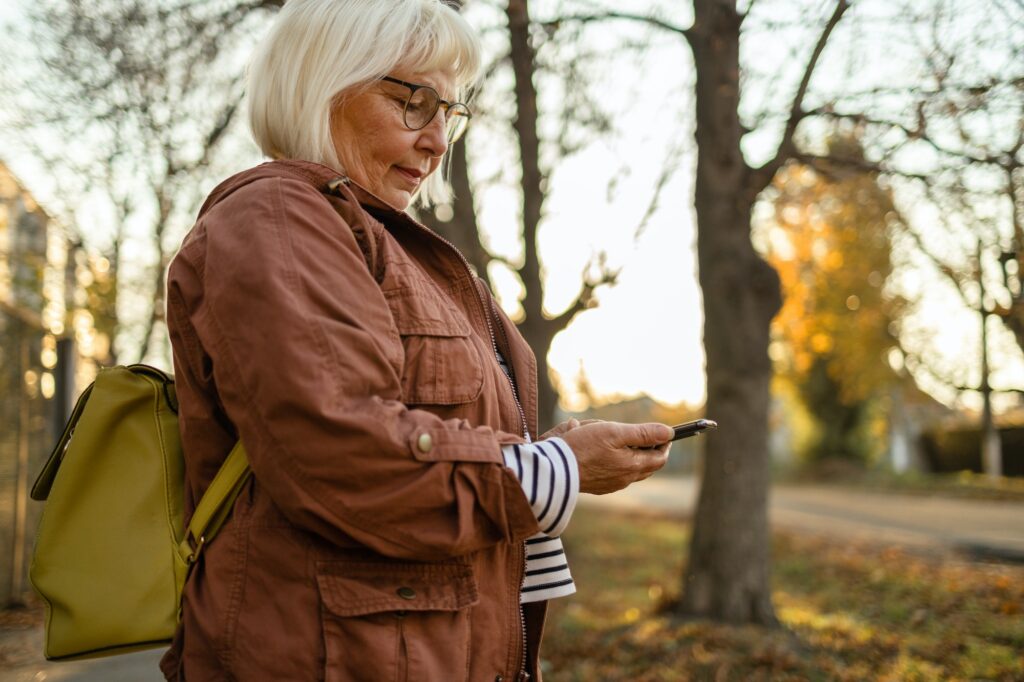 50s woman hand holding mobile phone checks messages and reads news in autumn nature at sunset