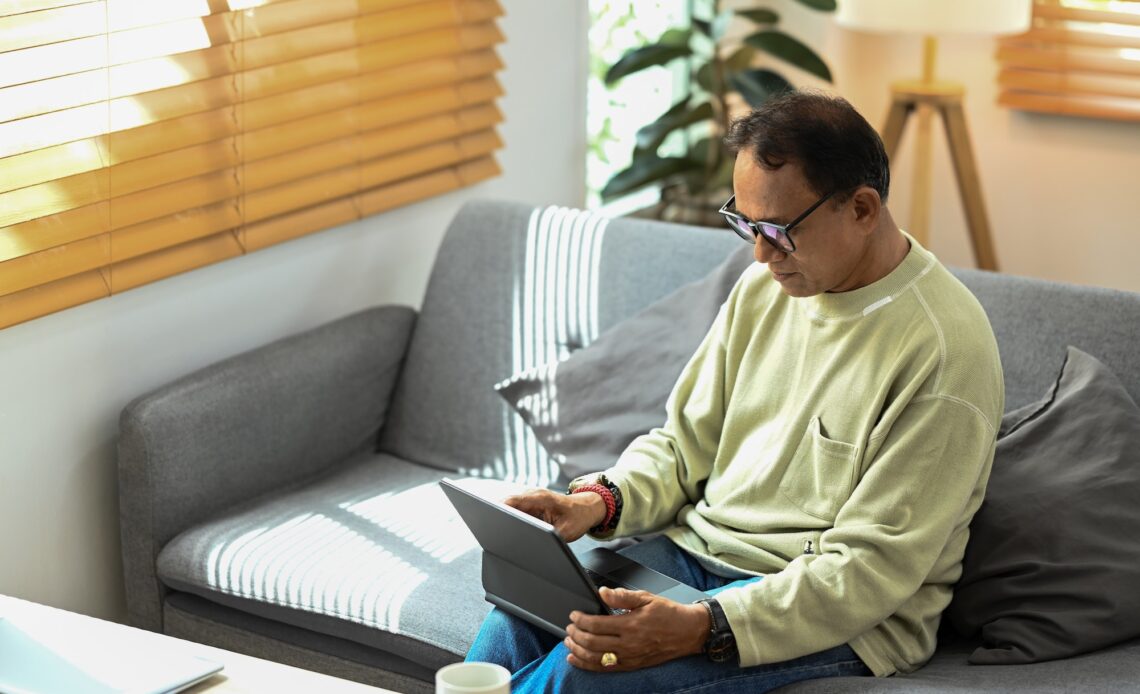 Mature man wearing eyeglasses reading online news on digital tablet while relaxing on couch.