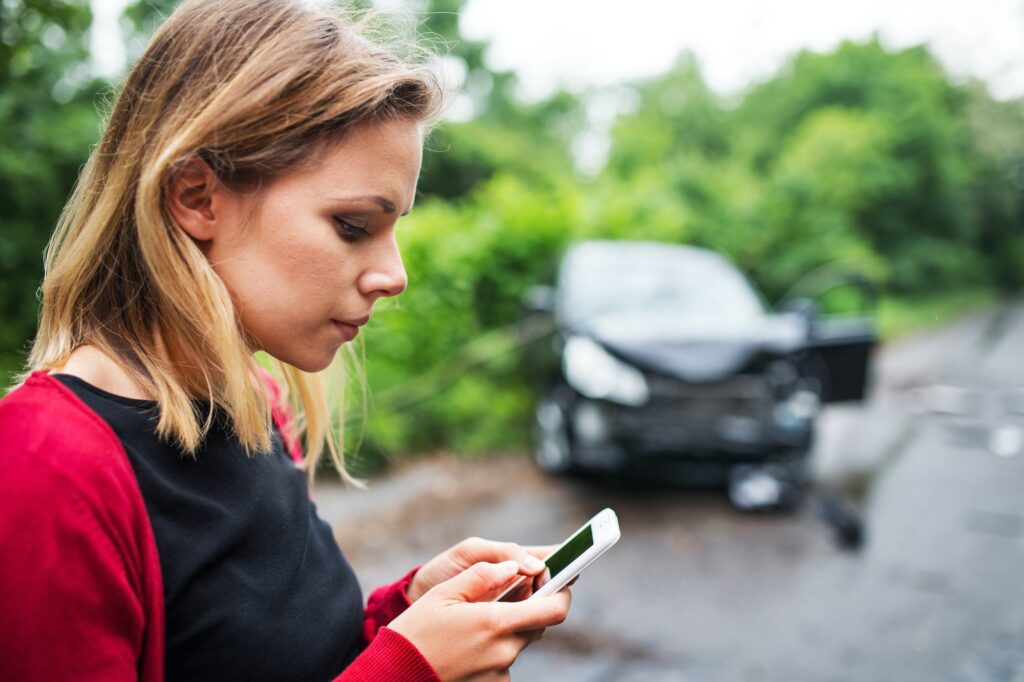 A young woman with smartphone by the damaged car after a car accident, text messaging.