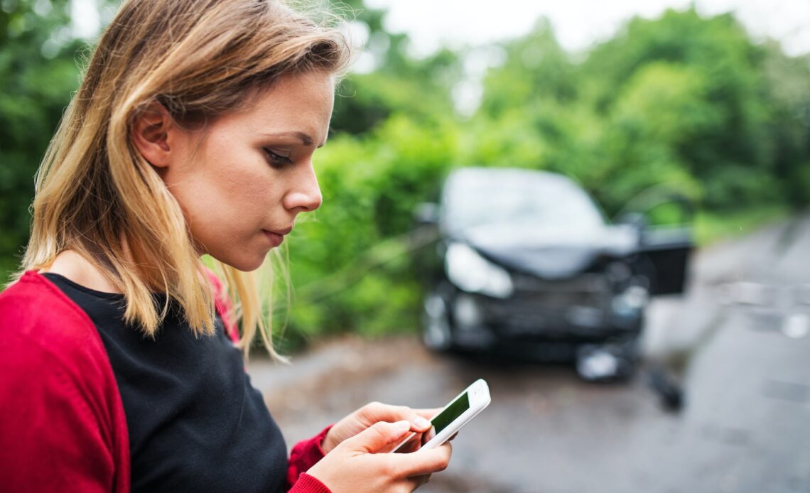 A young woman with smartphone by the damaged car after a car accident, text messaging.
